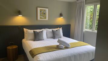 A neatly made bed with white linens and a yellow throw blanket, two wall-mounted lamps, a framed artwork, and a window with curtains.