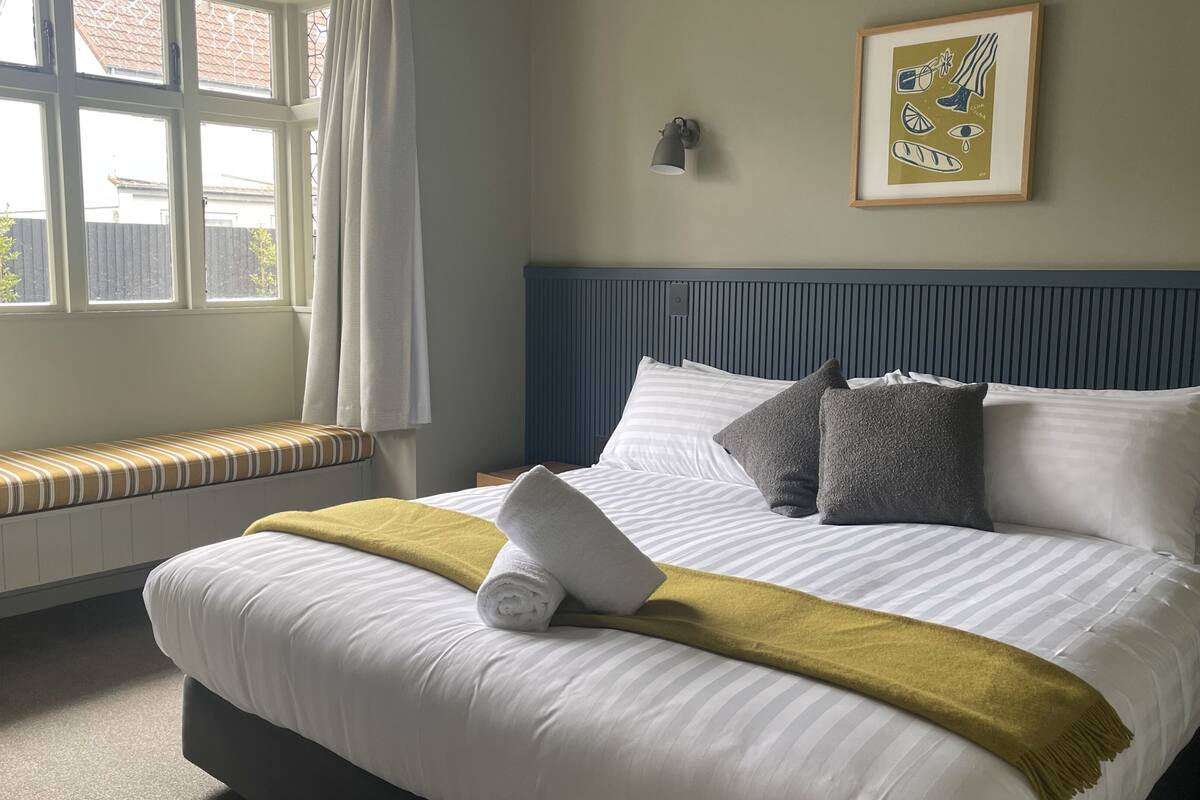 Een netjes opgemaakt bed, een opgerolde handdoek, een bankje bij het raam en een lijst met een afbeelding aan de muur.