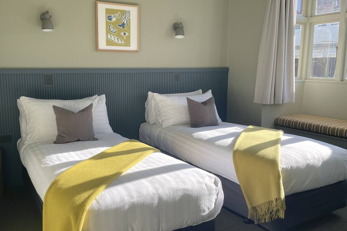 Een hotelkamer met twee bedden, een lijst met een kunstwerk aan de muur en een raam met gordijnen.