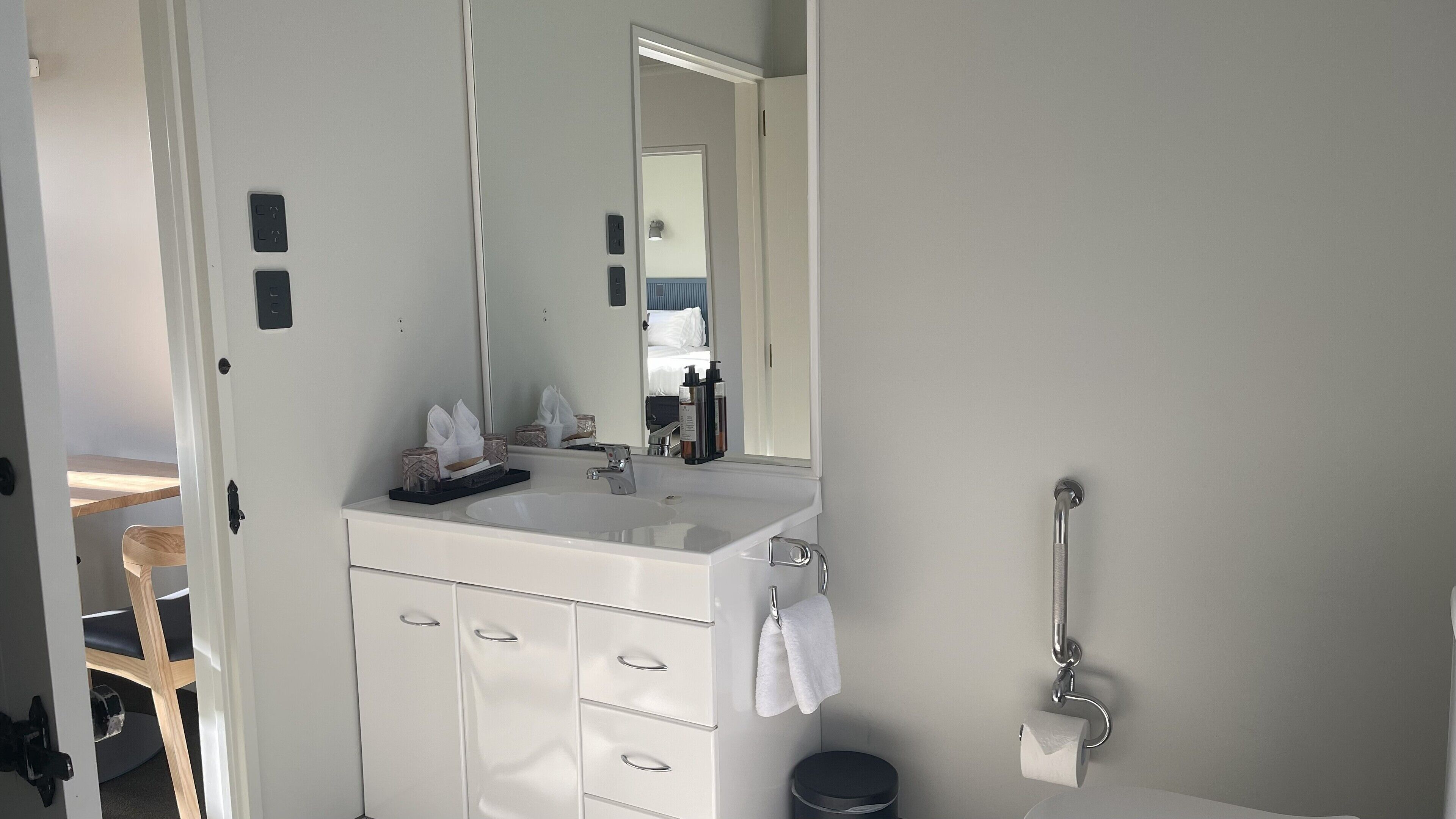 Un baño moderno con un lavamanos blanco, espejo y perchero. Se observa una silla de madera y un pequeño cesto de basura.