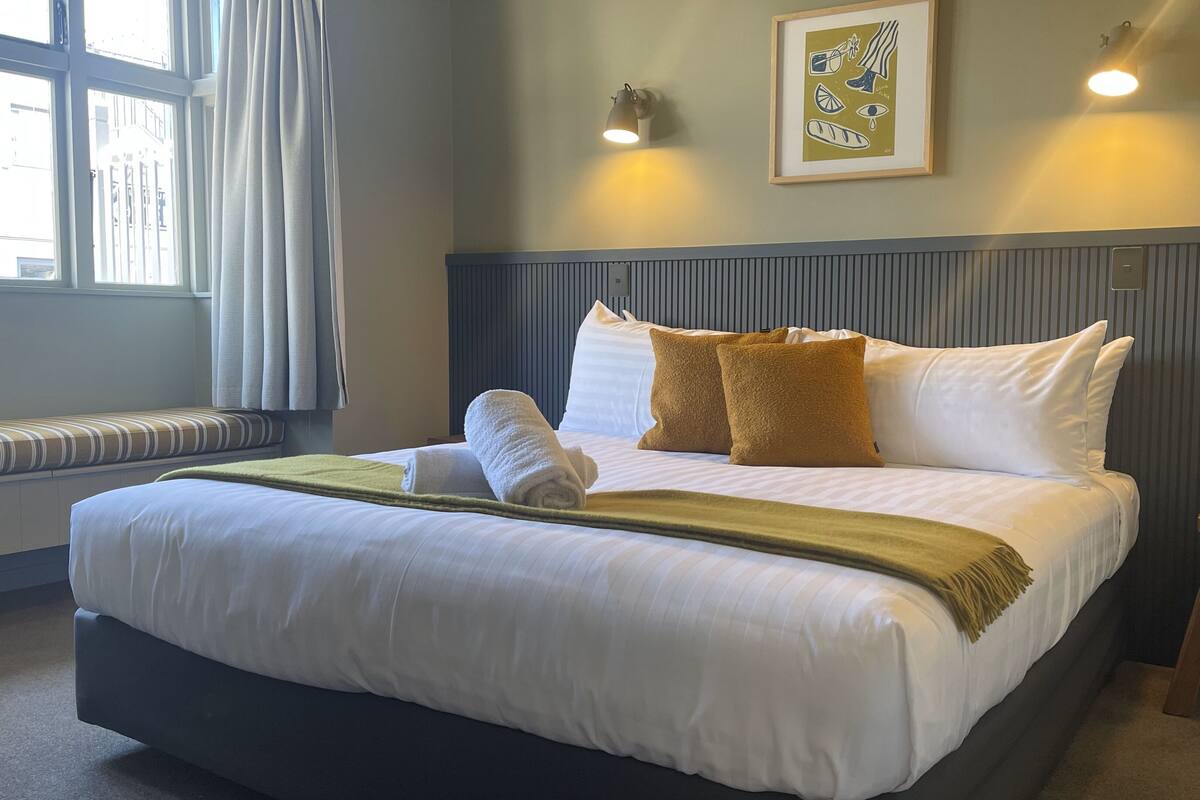 Een netjes opgemaakt bed met witte lakens, een groene plaid en twee bruine kussens. Boven het bed hangt een ingelijste foto, en links bevindt zich een raam met gordijnen.