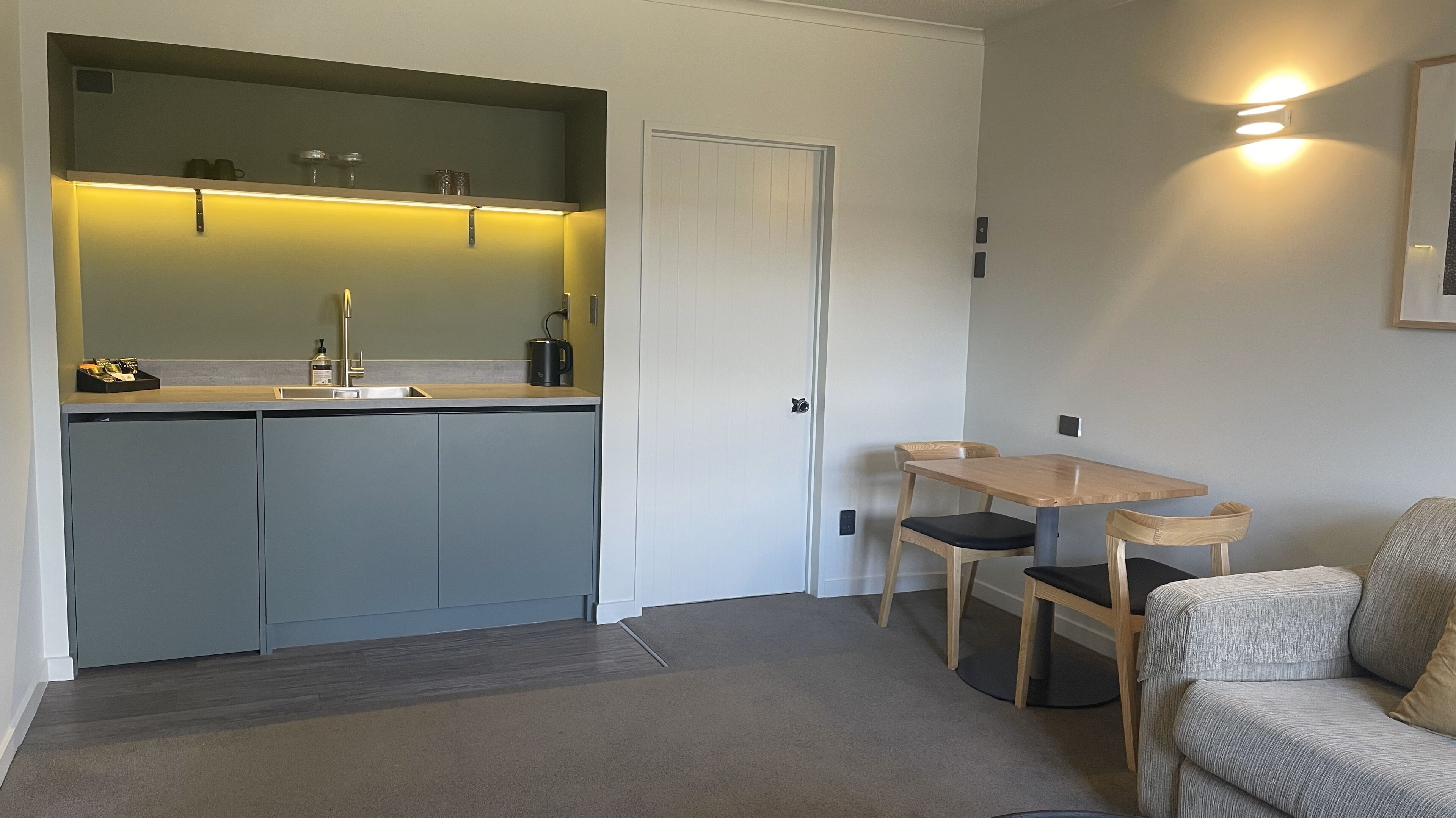 A compact kitchen with a sink, cabinets, and a countertop. There is a dining area with a table and chairs. A door is visible in the background.