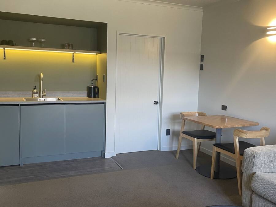 A compact kitchen with a sink, cabinets, and a countertop. There is a dining area with a table and chairs. A door is visible in the background.