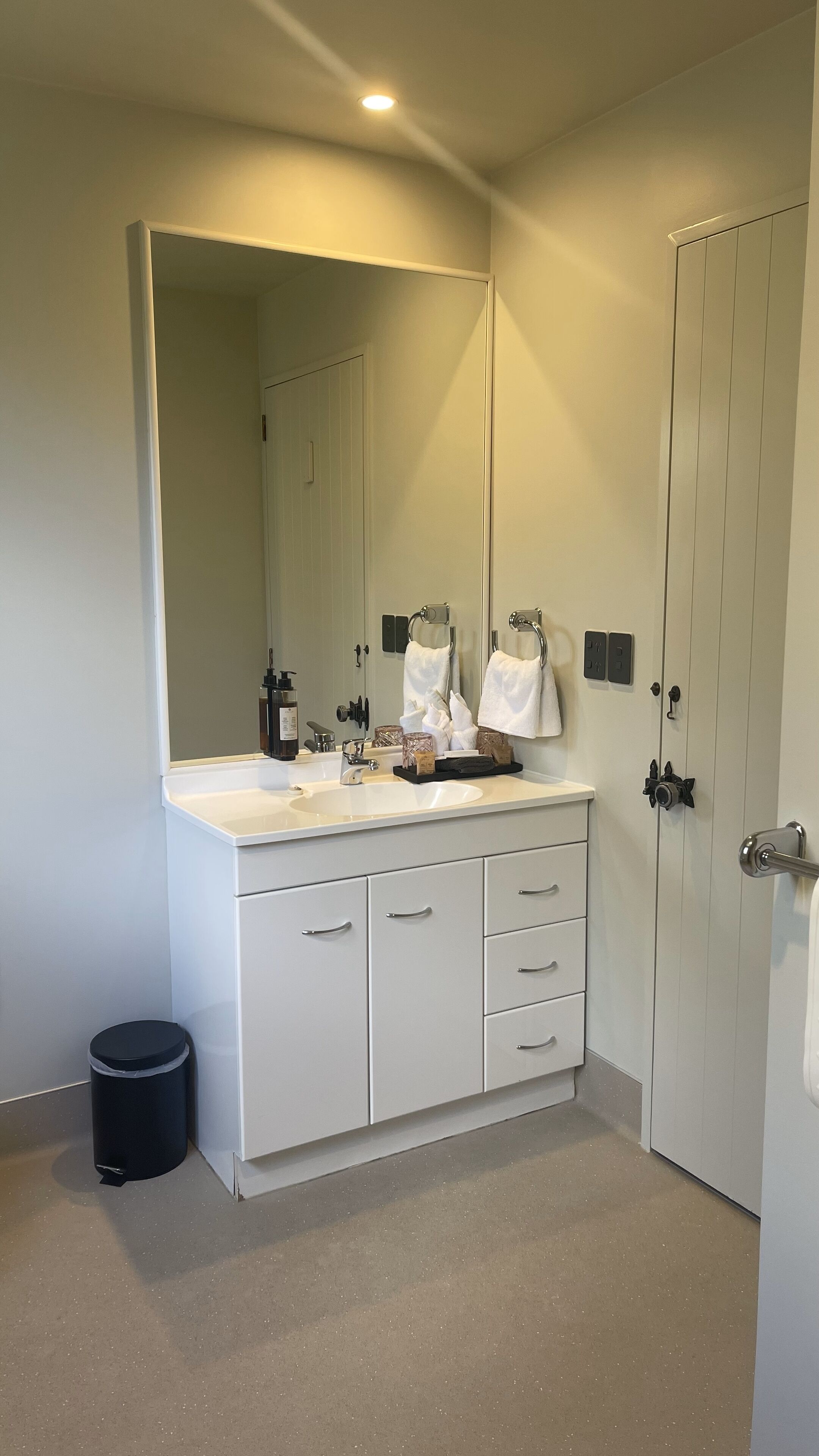 Et badeværelse med et hvidt toiletbord, et stort spejl, en vask og en sort skraldespand.