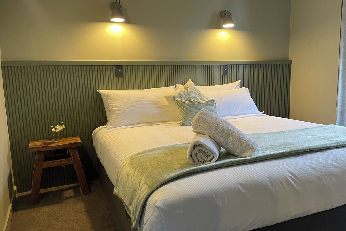 Een net opgemaakt bed met witte lakens, een opgerolde handdoek en een vaas met bloemen op een klein houten nachtkastje.