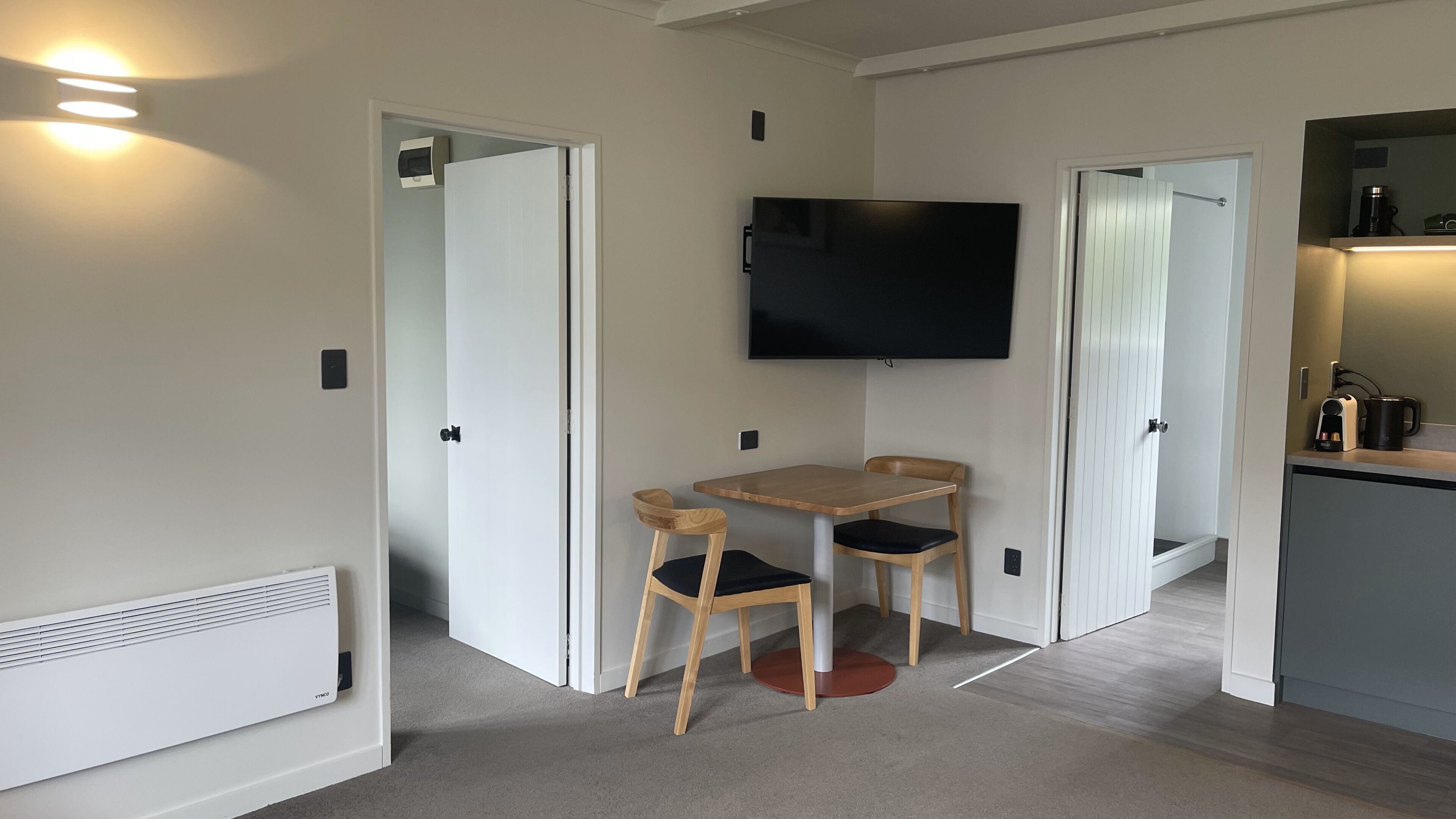 A compact room with a flat-screen TV mounted on the wall, a small wooden table with two chairs, and a kitchenette area with a coffee maker.