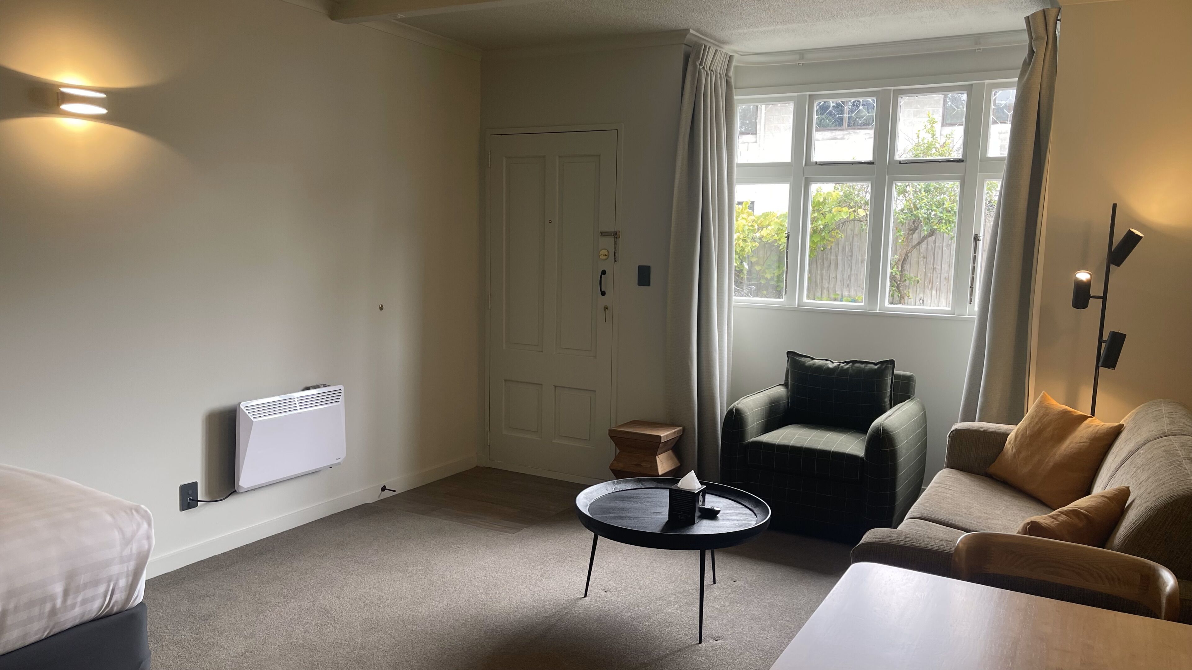 A living room with a white radiator, a round table with a black base, a dark armchair, and a beige sofa with a brown cushion.