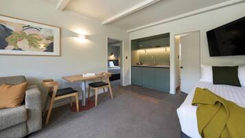 A hotel room with a bed, a sofa, a dining table with chairs, a kitchen area, and a TV mounted on the wall.