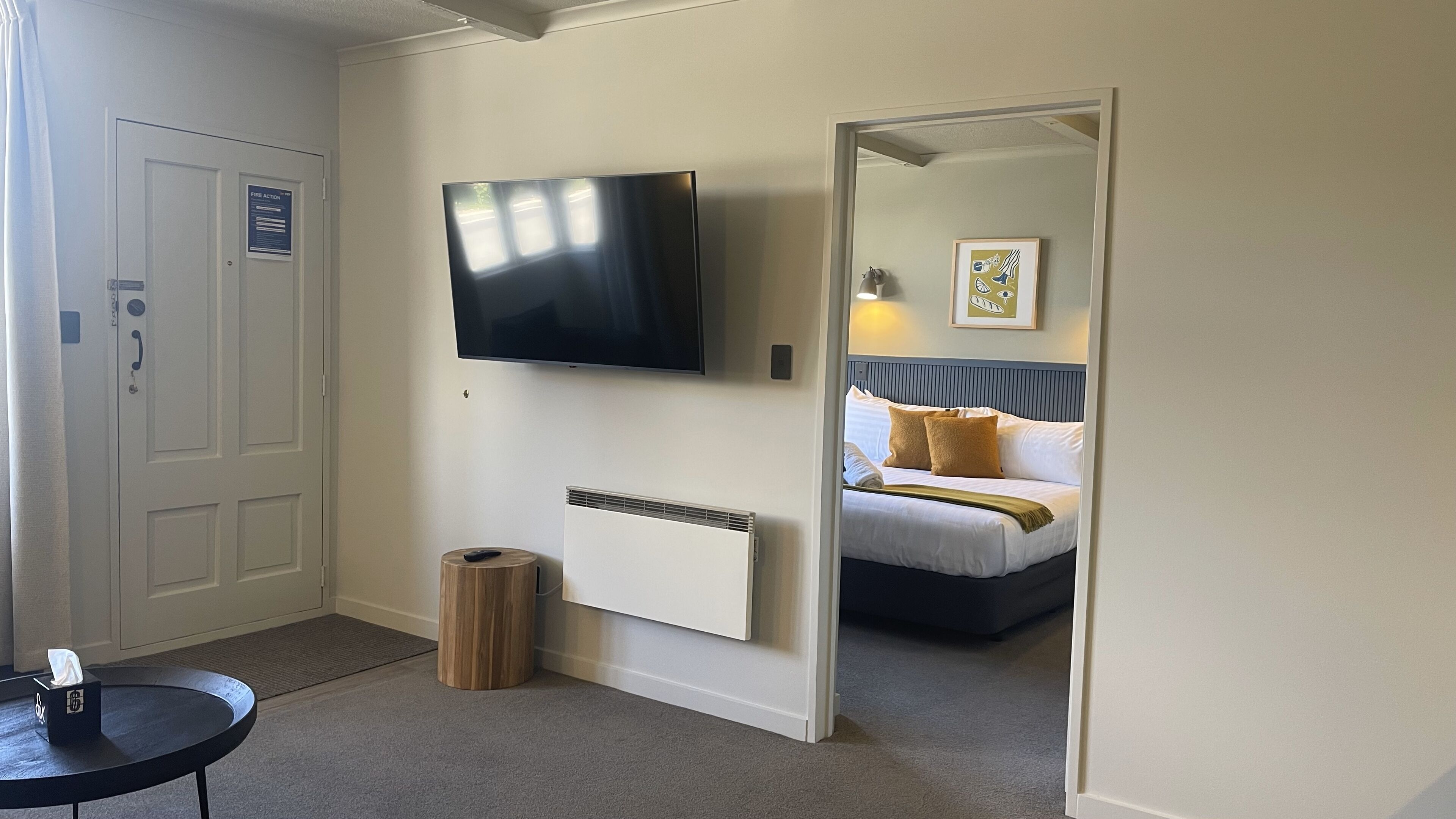 Habitación de hotel con cama, televisor montado en la pared, mesita de noche, cubo de basura y puerta con señal de alarma contra incendios.