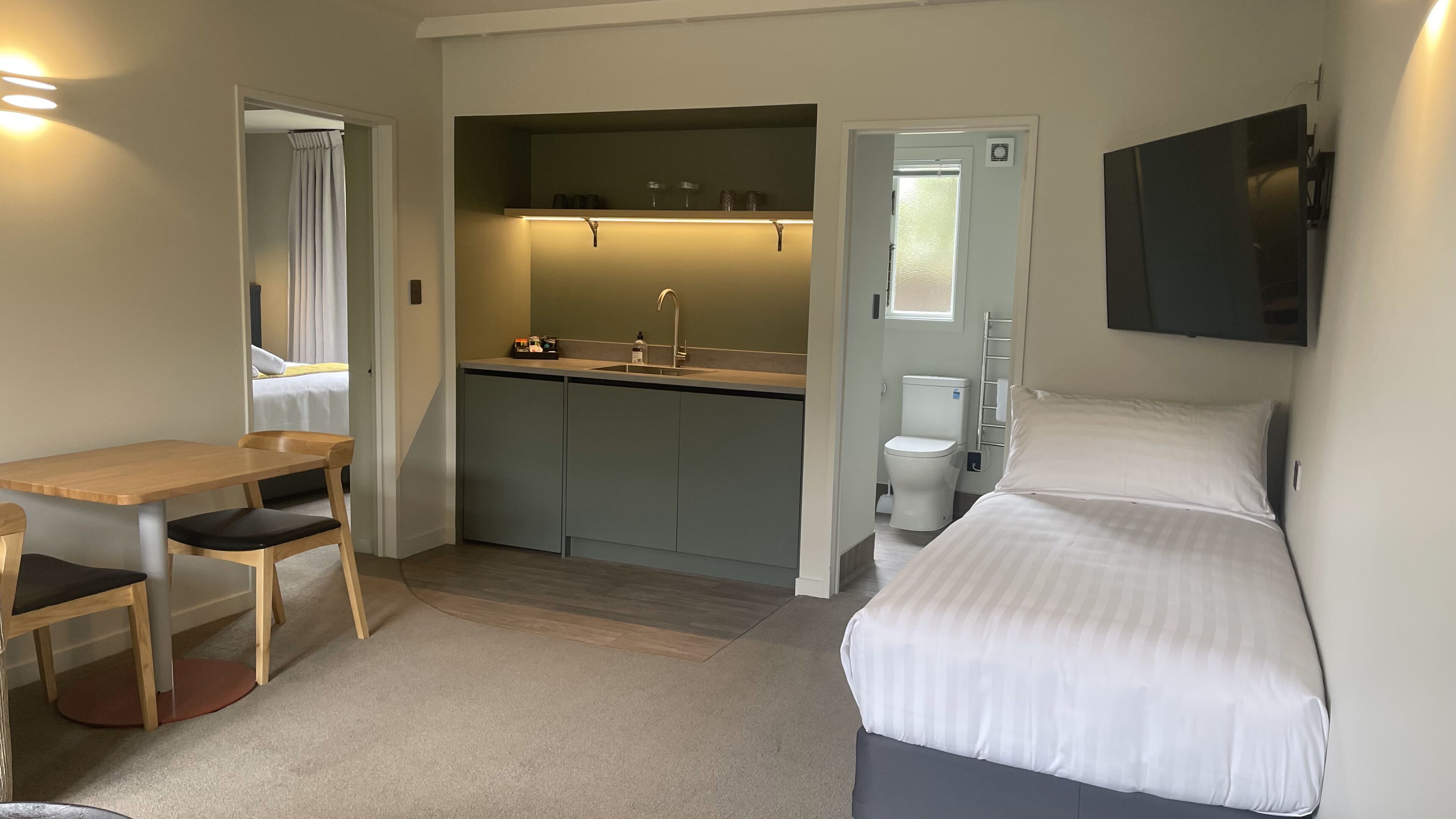 A compact hotel room with a bed, a desk, a chair, a kitchenette, and a bathroom.