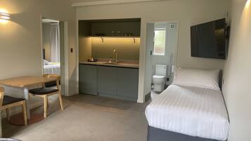 A compact hotel room with a bed, a desk, a chair, a kitchenette, and a bathroom.