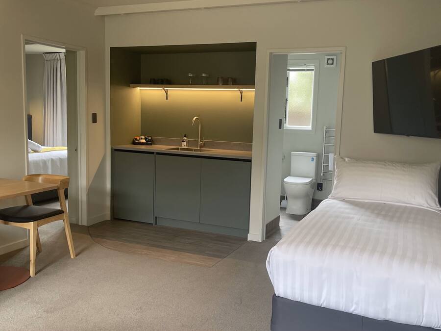 A compact hotel room with a bed, a desk, a chair, a kitchenette, and a bathroom.