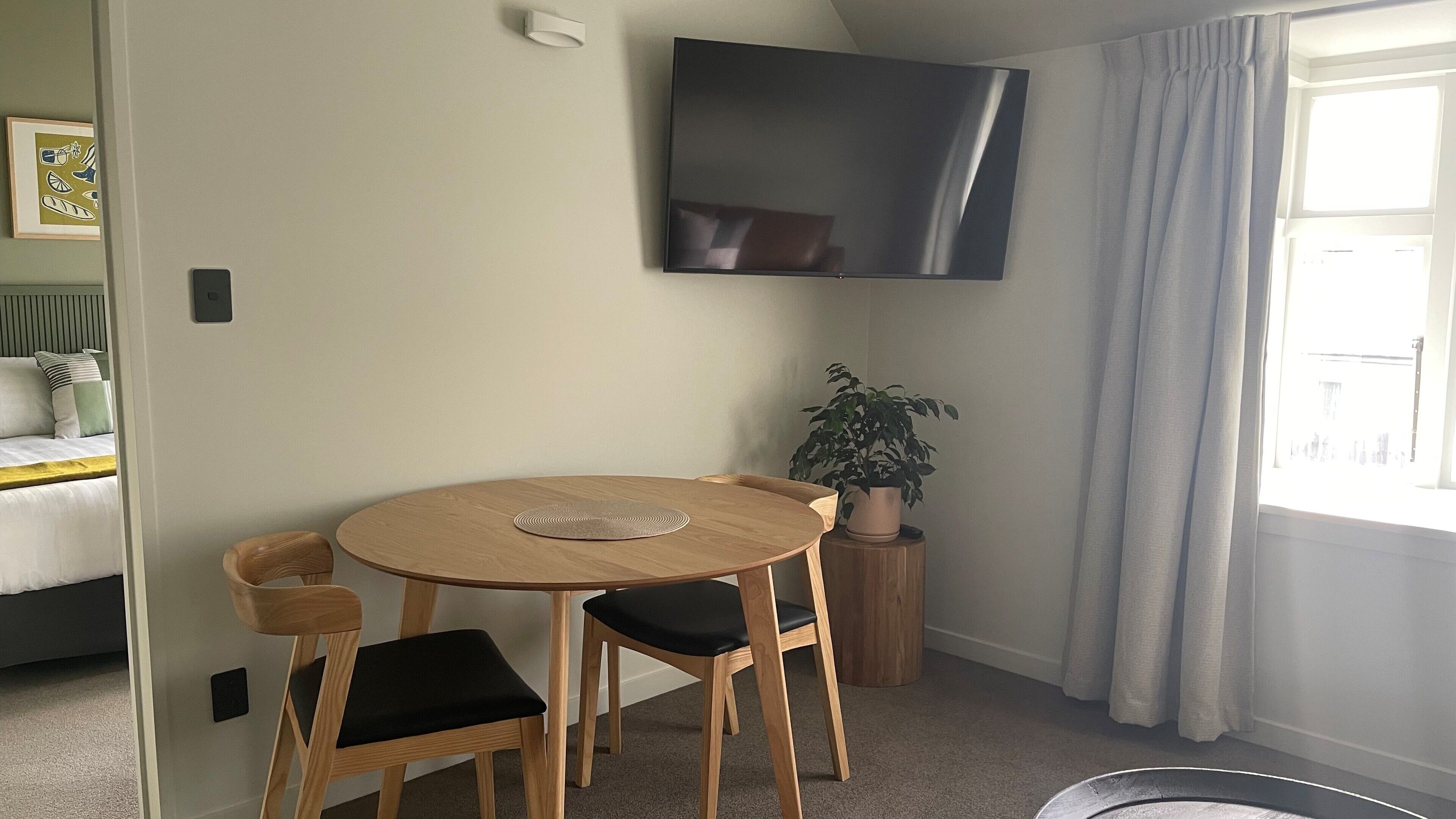A room with a round wooden table, two chairs, a potted plant, and a flat-screen TV mounted on the wall.