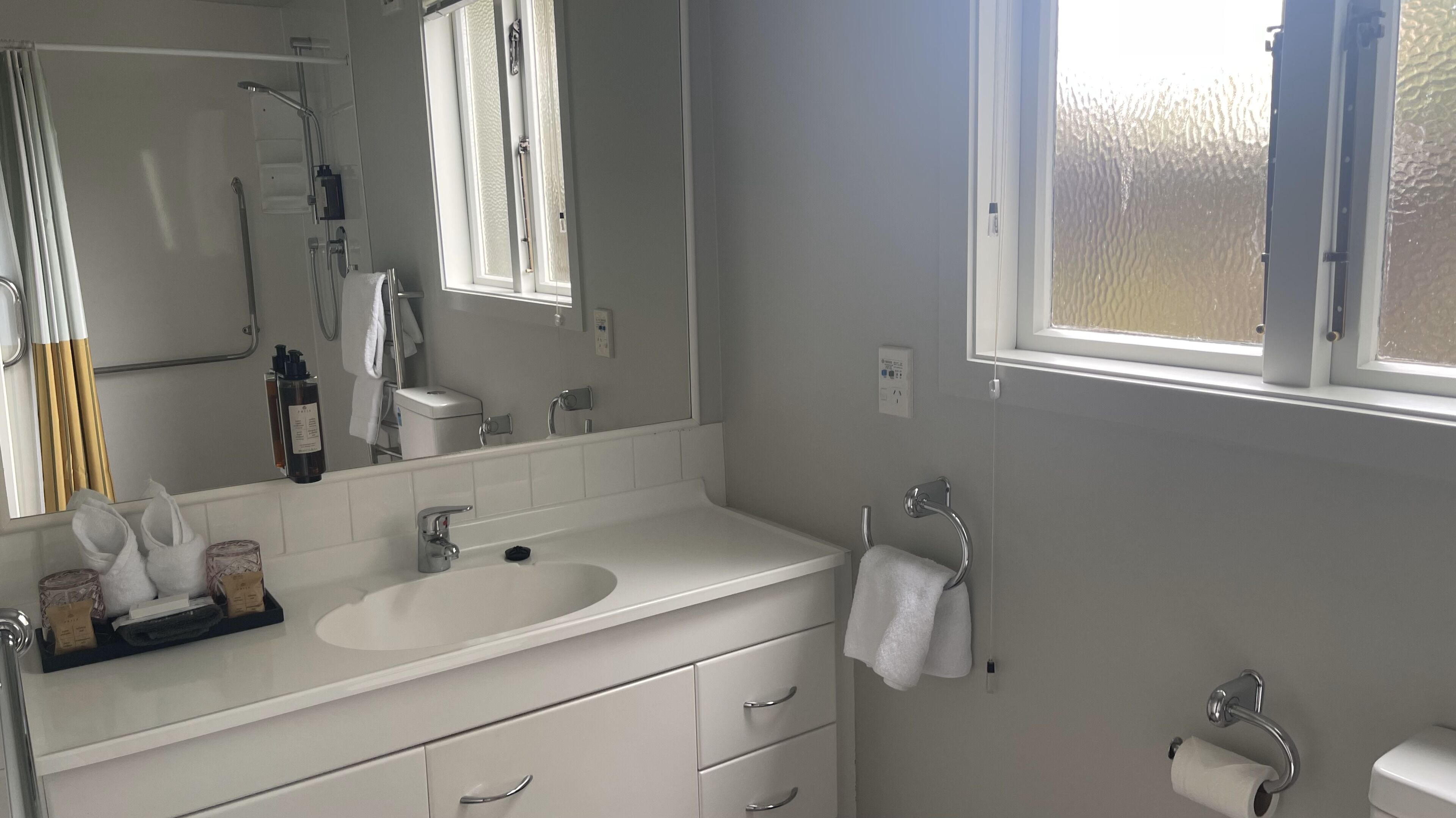 Une salle de bain moderne avec un lavabo blanc, un miroir et un meuble-lavabo. On y trouve également un porte-serviettes, des toilettes et une fenêtre qui laisse entrer la lumière naturelle.