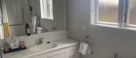 Une salle de bain moderne avec un lavabo blanc, un miroir et un meuble-lavabo. On y trouve également un porte-serviettes, des toilettes et une fenêtre qui laisse entrer la lumière naturelle.