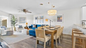 A modern living room with a blue sofa, wooden dining table, and a ceiling fan. - NEW! Oceanfront! Beach Vacation Bliss! 50 Steps ToThe Sand, Pool, Great Views. (Oak Island)