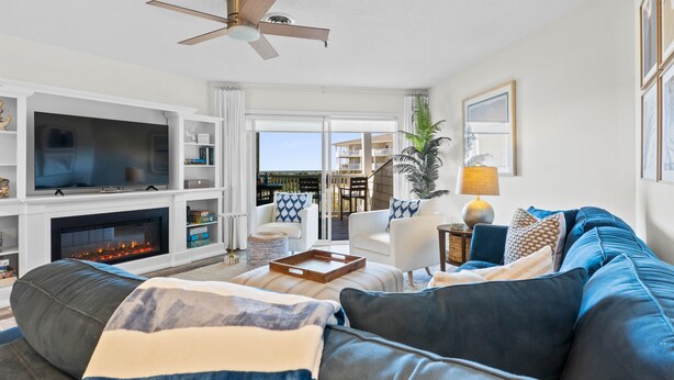 A living room with a fireplace, a large TV, a ceiling fan, and a view of the outdoors. - NEW! Oceanfront! Beach Vacation Bliss! 50 Steps ToThe Sand, Pool, Great Views. (Oak Island)