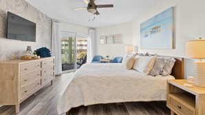 A bedroom with a large bed, a ceiling fan, a TV, a wooden dresser, a desk with a lamp, and a painting on the wall. - NEW! Oceanfront! Beach Vacation Bliss! 50 Steps ToThe Sand, Pool, Great Views. (Oak Island)