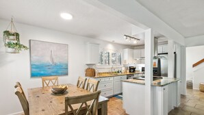 A modern kitchen with white cabinets, a wooden dining table, and a painting of a sailboat. - Dogs Welcome! Pool access. 1 Block to beach! Serene retreat under a majestic old oak canopy (Fernandina Beach)