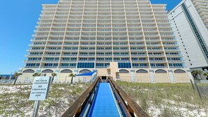 Un hotel grande con varios pisos y balcones, situado cerca de una playa con un paseo marítimo azul que conduce a la entrada.