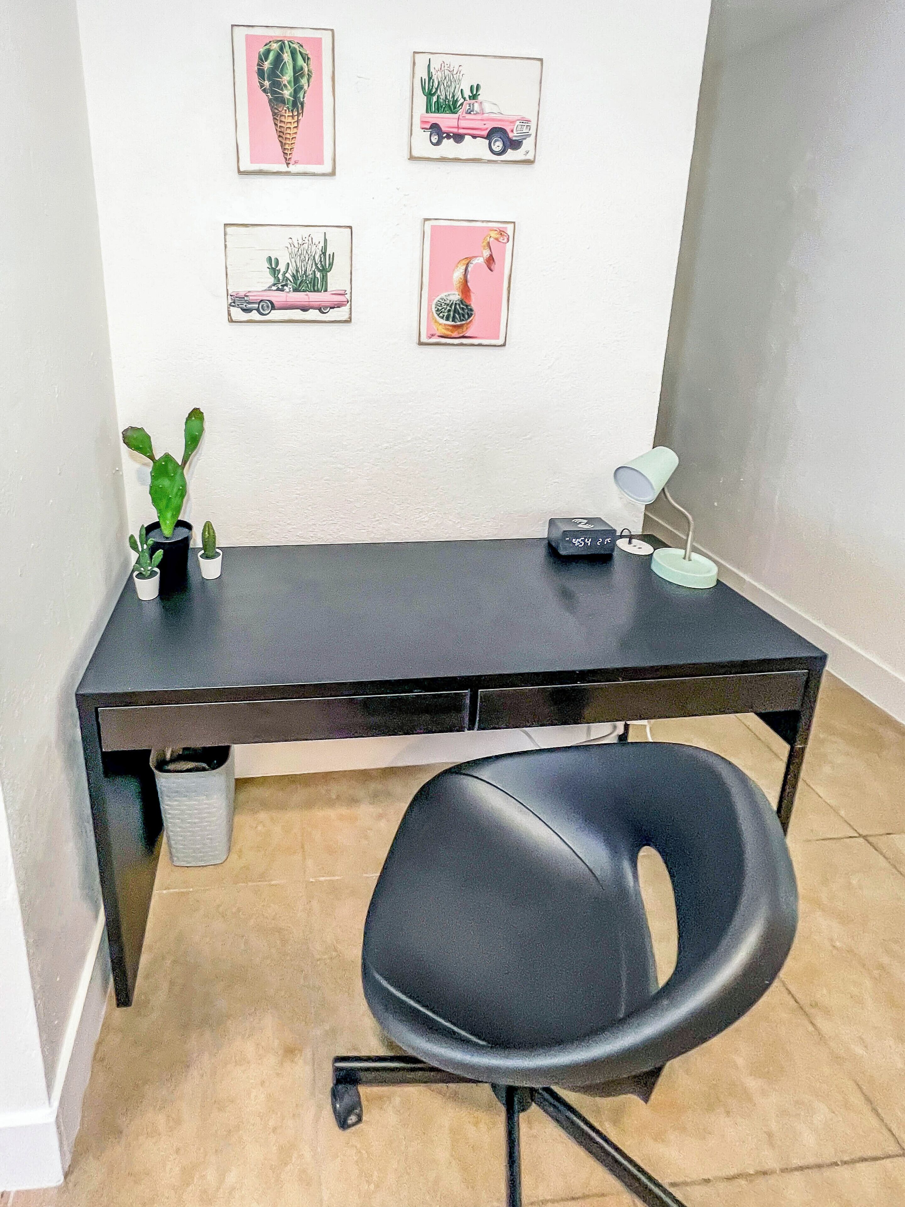 A dark wooden desk with a black office chair, a small cactus plant, and a desk lamp. There are four framed pictures on the wall above the desk.