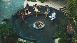 Un espacio de estar al aire libre circular con un brasero, rodeado de plantas y una estructura con techo de paja.
