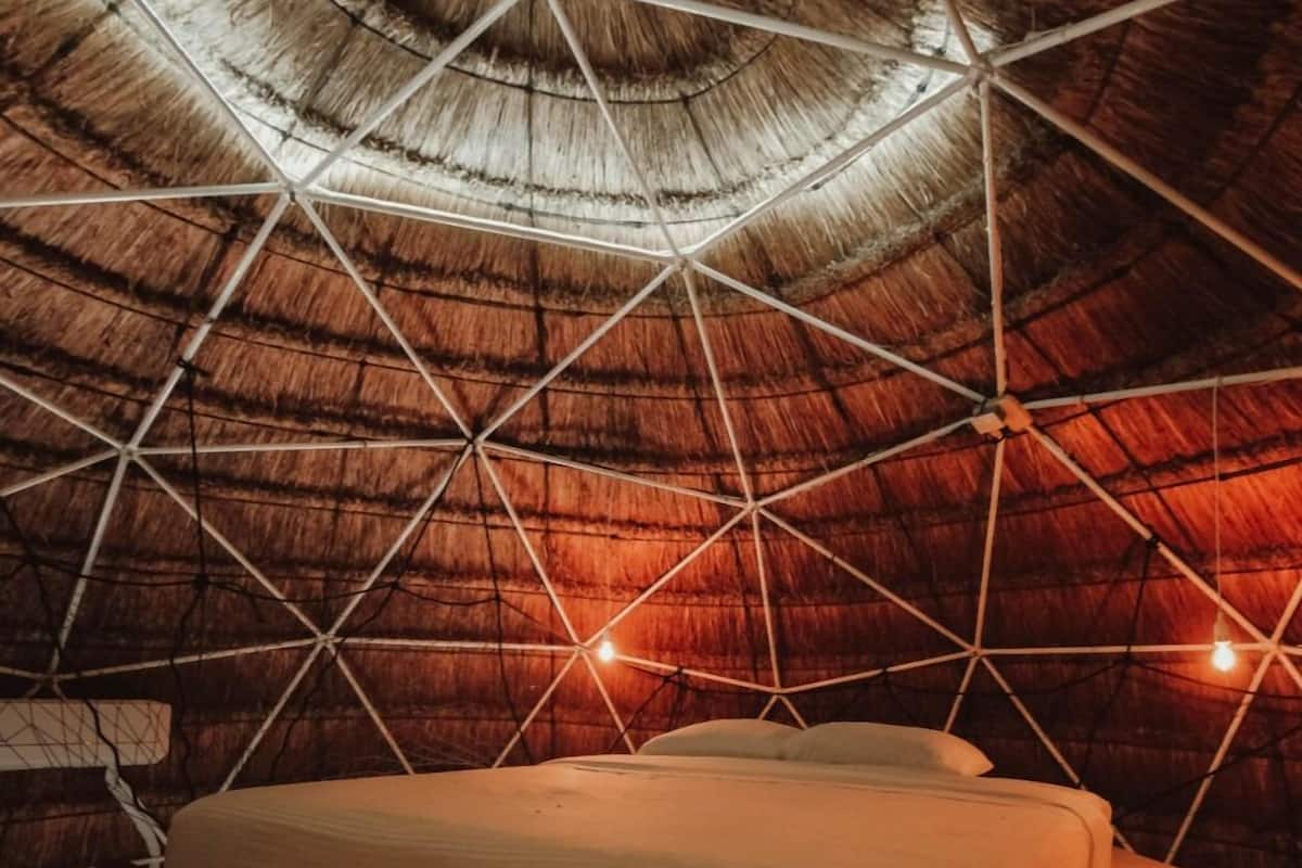 Quarto com telhado de colmo e estrutura metálica geométrica. Há uma cama com roupa de cama branca.