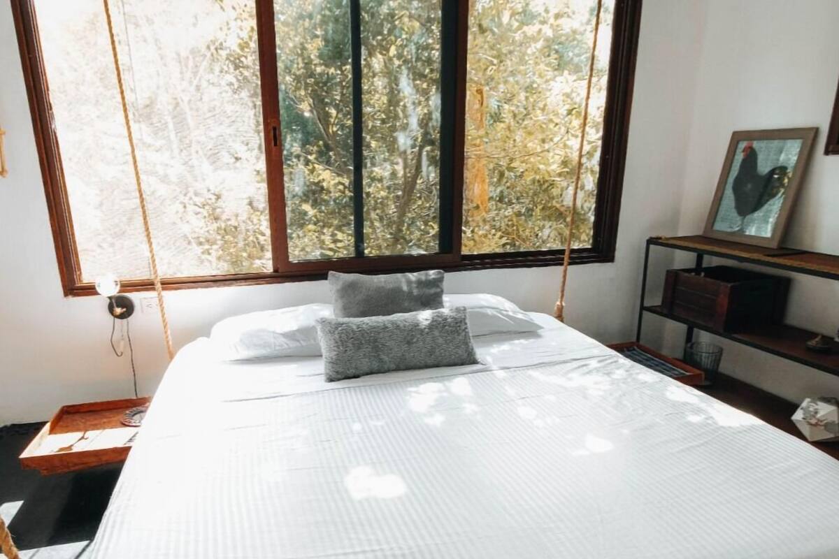 Quarto com uma janela grande, uma cama com roupa de cama branca, um travesseiro cinzento e uma mesa de cabeceira de madeira com uma moldura.