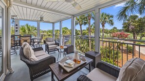 A covered outdoor area with wicker furniture, a table with books and drinks, and a view of palm trees and greenery. - PERFECT 2BR Seabrook Getaway, Pool Access, Golf View, One Block to Beach & Club! (Seabrook Island)