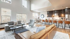 Un grand espace de vie comprenant un canapé, un fauteuil et une table basse, un coin repas avec des chaises, et une cuisine avec des tabourets de bar.