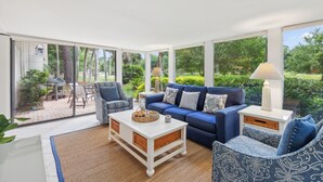Een ruime woonkamer met een blauwe bank, gestreept fauteuils en een witte salontafel. Buiten de schuifpui is een barbecue zichtbaar.