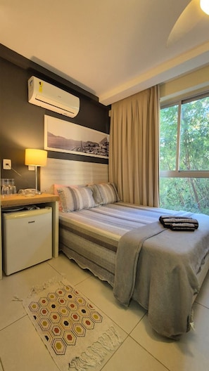 Habitación de hotel con cama, escritorio con lámpara, refrigerador pequeño, ventana con cortinas y aire acondicionado de pared.