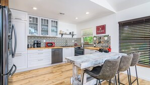 A modern kitchen with white cabinets, a wooden countertop, a stainless steel refrigerator, a red microwave, and a dining area with a wooden table and bar stools. - Surf House Cocoa Beach...Family-Friendly, Walk to Beach! (Cocoa Beach)