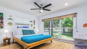 A bedroom with a wooden bed, a ceiling fan, a window with a view of greenery, and wall art. - Surf House Cocoa Beach...Family-Friendly, Walk to Beach! (Cocoa Beach)