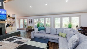 A spacious living room with a large gray sectional sofa, a black coffee table, and a fireplace. There are large windows providing natural light and a view of greenery outside. - Peaceful Retreat near Texas Tech with Epic Game room! (Lubbock)