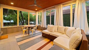 A modern living room with a wooden ceiling, a ceiling fan, a sofa, a dining table with chairs, and large windows with curtains.