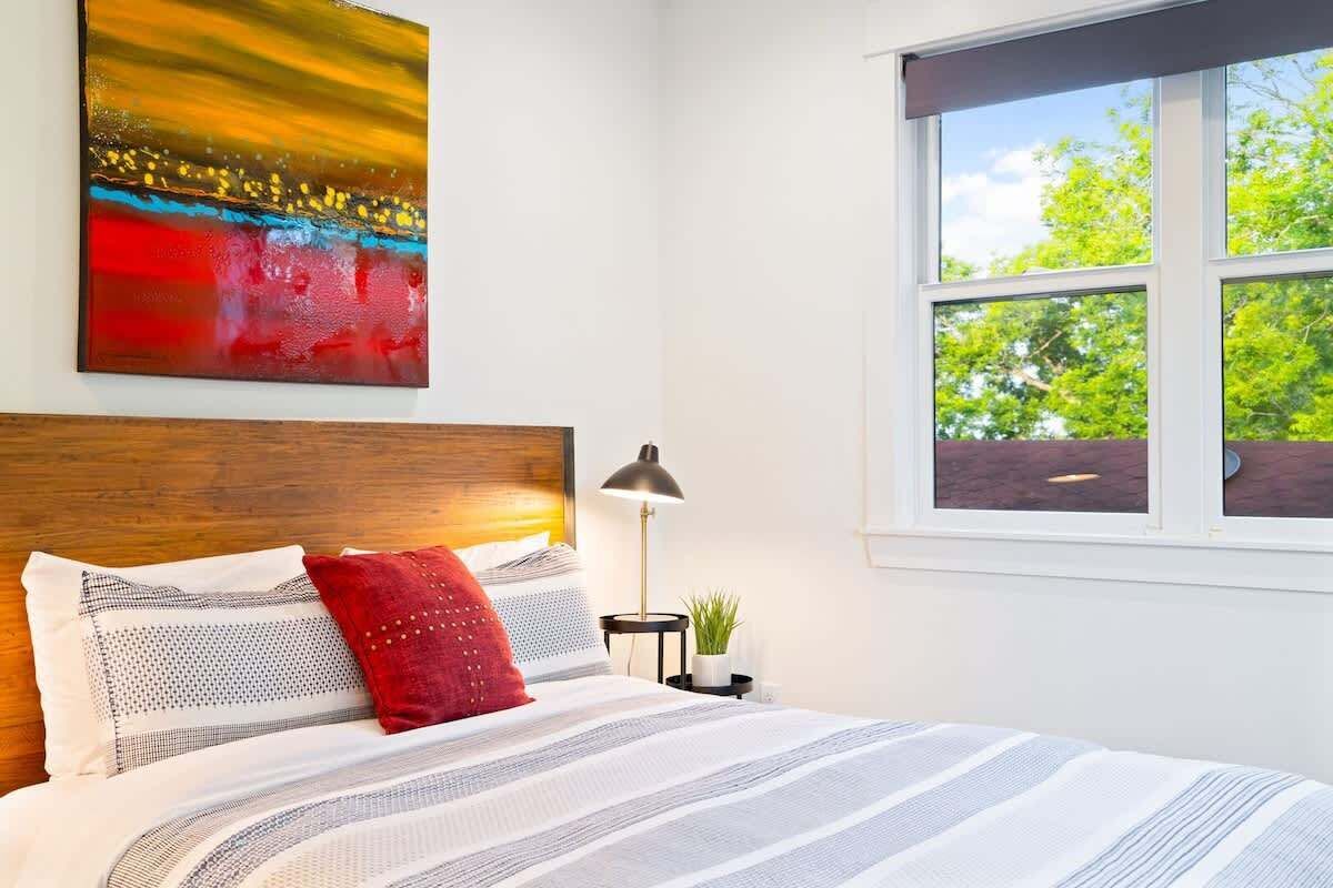 A bedroom with a wooden headboard, a bed with striped bedding, a red pillow, a nightstand with a lamp, and a window with a view of trees.