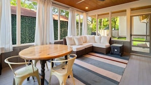 A modern living room with a wooden ceiling, a round wooden table with two chairs, a sectional sofa, and a large window with curtains.