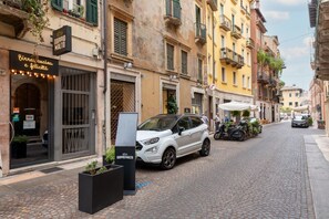 Uma rua de paralelepípedos com uma SUV branca estacionada na lateral, um edifício com uma placa indicando 'DOPPIO MALTO' e outro edifício com uma placa 'Biree, cucina e felicita'.