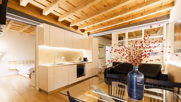 A modern kitchen with a glass dining table, wooden ceiling beams, and a vase with flowers.