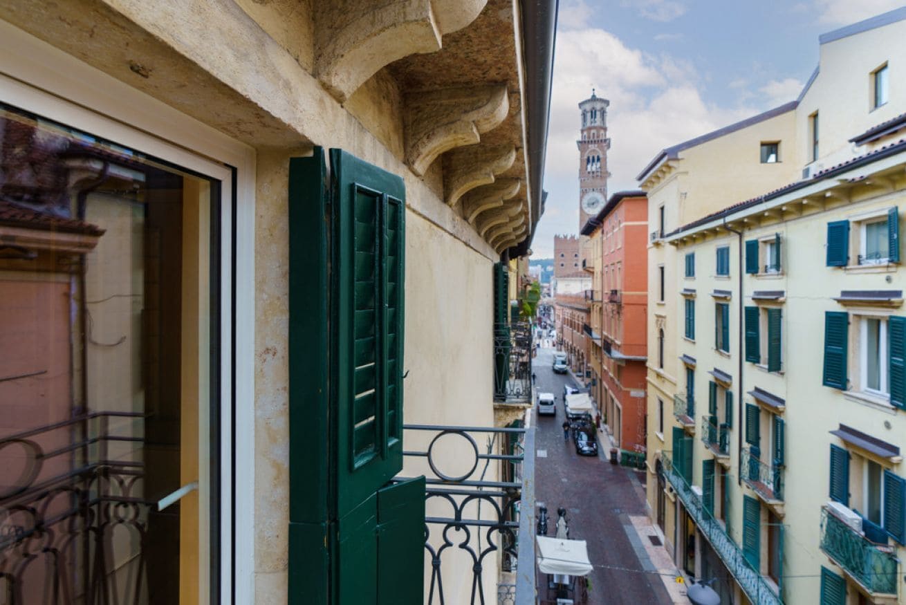 Une rue étroite, bordée de bâtiments des deux côtés, un balcon avec des volets verts et, au loin, un clocher.