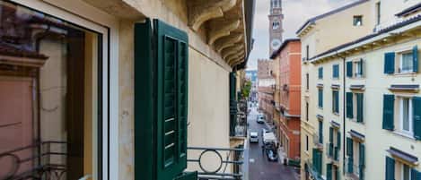 A narrow street with buildings on either side, a balcony with green shutters, and a clock tower in the distance.