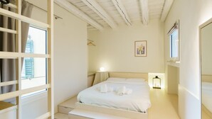 Quarto com cama, mesas de cabeceira, um criado-mudo, janela com cortinas, quadro na parede e teto com vigas aparentes.