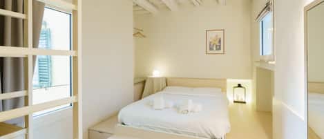 A bedroom with a bed, bedside tables, a nightstand, a window with curtains, a wall-mounted picture, and a ceiling with exposed beams.