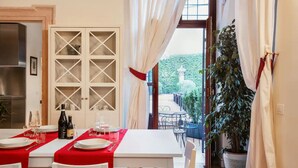 Un comedor con una mesa dispuesta para dos personas, una botella de vino y copas. Hay un armario blanco con puertas de cristal y se ve un jardín a través de una puerta abierta.