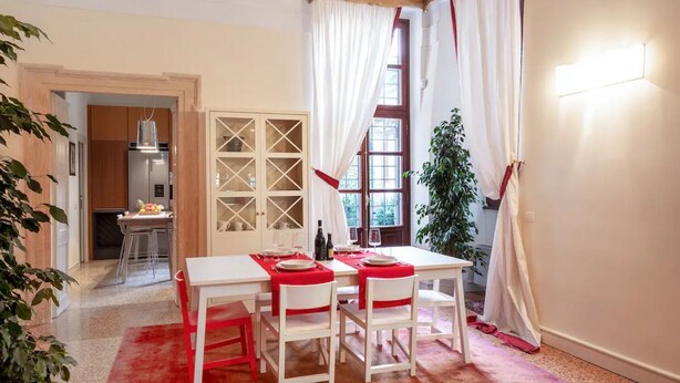 Un comedor con una mesa dispuesta para cuatro, una cocina al fondo y un gran ventanal con cortinas.