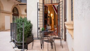 Un área de estar al aire libre con mesa y sillas, una planta en maceta y vista hacia la entrada de un edificio.