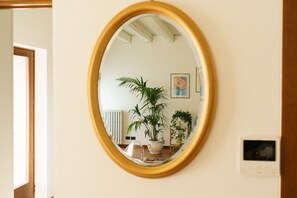 Um espelho oval fixado na parede, com um radiador, um vaso de planta e uma pequena prateleira com um quadro.