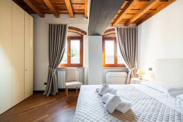 A bedroom with a bed, two windows with curtains, a chair, a bedside table with a lamp, and a radiator. - Merici residence, elegant and fairytale-like (Verona)