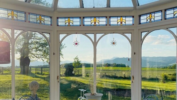 Une véranda dotée d’un toit en verre, offrant une vue sur un paysage verdoyant et des montagnes. À l’intérieur, on trouve une table avec des chaises et un vase décoratif.